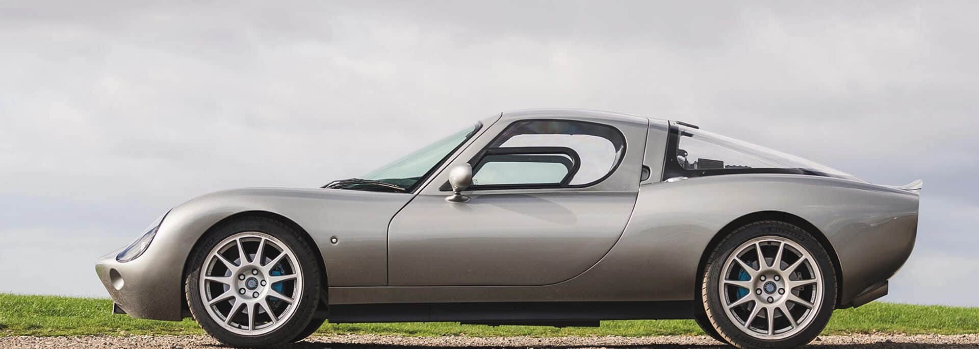 Silver Wells Motor Cars sports car with open door, displayed on gravel against green grass and cloudy sky background.
