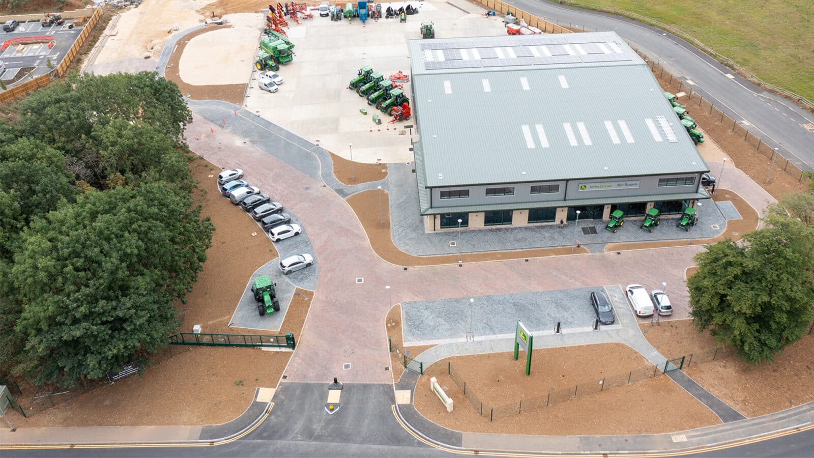 Agricultural facility at Oakham featuring modern shed with solar panels, green tractors, parking areas, and landscaped grounds surrounded by woodland.