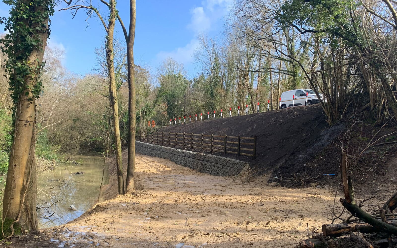 Landslide stabilisation site featuring wooden retaining wall, geotextile reinforcement, and drainage measures along wooded slope adjacent to watercourse.