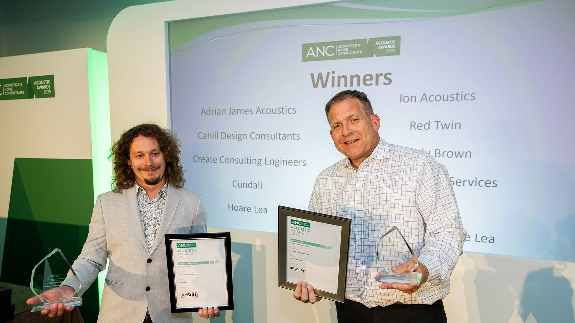 Two men stand holding framed certificates and glass awards in front of a projected winners list at an awards event.