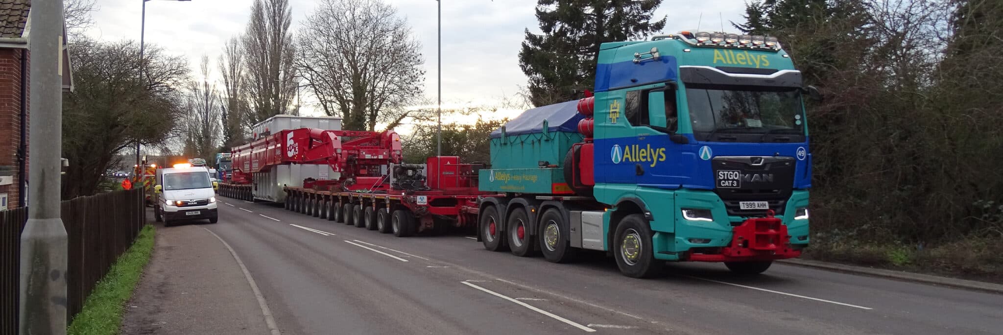 Abnormal load convoy on UK road transporting wind farm components for Hornsea 3 offshore wind farm, with Allelys haulage truck leading red agricultural trailers.