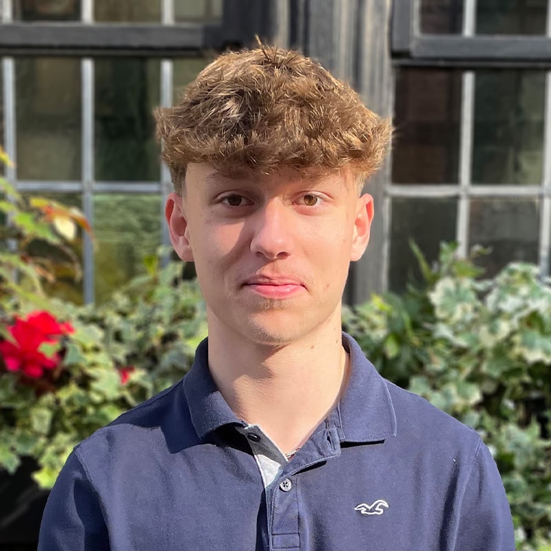 Louie, new starter at Create Consulting Engineers, posed outdoors against a greenhouse backdrop with flowering plants.
